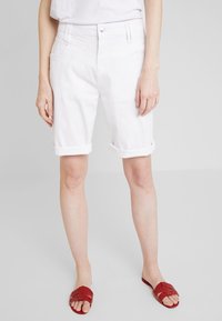 Shorts en denim blanc avec des ourlets roulés, design à cinq poches et fermeture par bouton supérieur. Texture lisse et coupe standard. Associés à des sandales à glisser rouges.