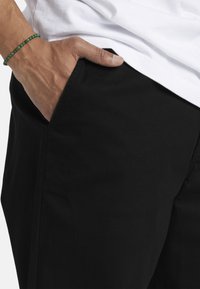Black pants made of smooth fabric, featuring a side pocket and a close-up of a hand resting on the fabric. White t-shirt partially visible.