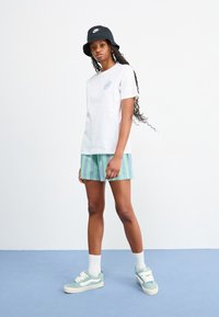 Hvit bomull T-skjorte med en lys blå sirkulær logo, grønne og blå stribede shorts, svart bøttehatt og mintgrønne sneakers med hvite detaljer.