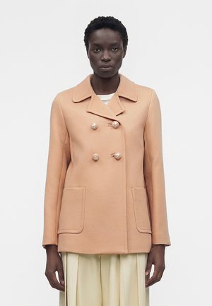 Άτομο με σκούρο δέρμα φοράει μπεζ διπλής σειράς παλτό με κουμπιά από πέρλες και μεγάλες μπροστινές τσέπες, στέκεται μπροστά σε μονόχρωμο φόντο.