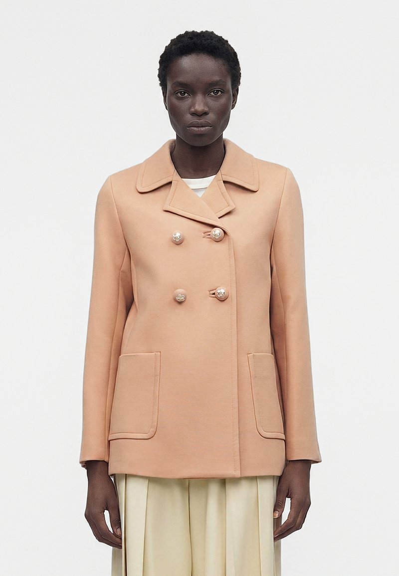 Άτομο με σκούρο δέρμα φοράει μπεζ διπλής σειράς παλτό με κουμπιά από πέρλες και μεγάλες μπροστινές τσέπες, στέκεται μπροστά σε μονόχρωμο φόντο.