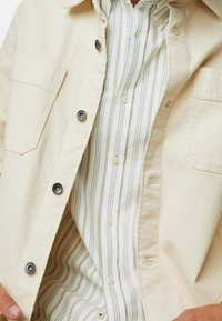 Chaqueta de mezclilla beige con botones negros, un bolsillo en el pecho, llevada sobre una camisa de botones a rayas blancas y grises con cuello estándar.