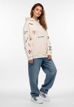 Sweatshirt à capuche couleur crème avec des impressions graphiques mélangées et du texte, associé à un jean bleu ample et des sneakers blanches avec des rayures noires.