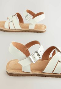 Sandales en cuir blanc avec des brides croisées, des boucles ajustables et une semelle naturelle de couleur tan. Semelle plate avec surface texturée et design minimal.