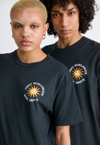 Svart bomull t-skjorter med et hvitt og oransje solgrafikk og teksten "OBEY WORLDWIDE RISE ABOVE". Korte ermer, rund halsdesign.
