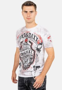 T-shirt bianca a maniche corte con grafica nera e rossa, caratterizzata da un design a scudo e dettagli infuocati. Tessuto morbido con un aspetto usurato.