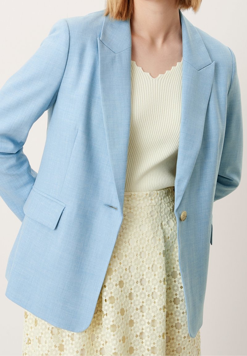 Femme portant un blazer bleu clair sur un haut côtelé crème et une jupe en dentelle crème, posant avec une main sur la hanche devant un fond uni.