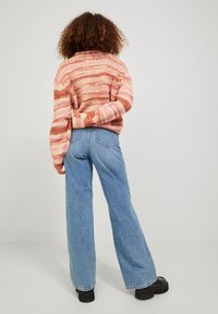 Svetr s dlouhým rukávem s růžovými a oranžovými pruhy, doplněný vysokými modrými džíny, nošený s černými robustními botami.