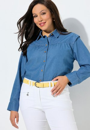 Donna con capelli lunghi e scuri, indossa una camicia blu con bottoni, pantaloni bianchi e una cintura gialla, sorridente con una mano sulla vita.