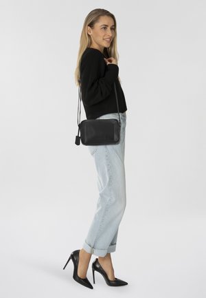 Borsa a tracolla in pelle nera con finitura texturizzata, chiusura con zip e tracolla regolabile, abbinata a jeans a righe blu chiaro e tacchi neri.