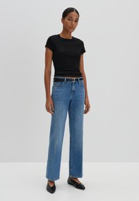 Zwart getailleerd T-shirt gecombineerd met hooglopende lichtblauwe wijde jeans, zwarte riem en zwarte puntige ballerina's op een effen achtergrond.