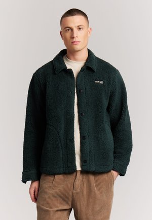 Junger Mann mit kurzen Haaren, der eine grüne Fleecejacke über einem weißen Hemd und braunen Cordhosen trägt, steht vor einem einfarbigen Hintergrund.