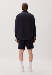 Marineblauwe longsleeve shirt met omgeslagen mouwen, gecombineerd met marineblauwe shorts. Het model draagt witte sneakers en sokken, wat een minimalistische stijl uitstraalt.