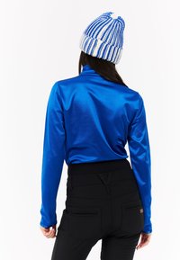 Modrý lesklý top s dlouhým rukávem a vysokým límcem, který je spárován s černými kalhotami s zadními kapsami a strukturovaným pasem, nošený s pruhovanou pletenou čepicí.