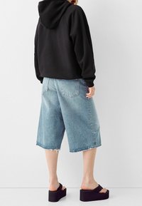 Sudadera corta negra, culottes de mezclilla azul claro con dobladillo deshilachado y sandalias de plataforma negras. Presenta una capucha y bolsillos laterales.