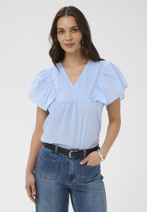 Femme aux longs cheveux foncés portant un chemisier bleu clair à col en V avec des manches à volants, un jean bleu, une ceinture noire et un bracelet argenté, debout devant un fond blanc.