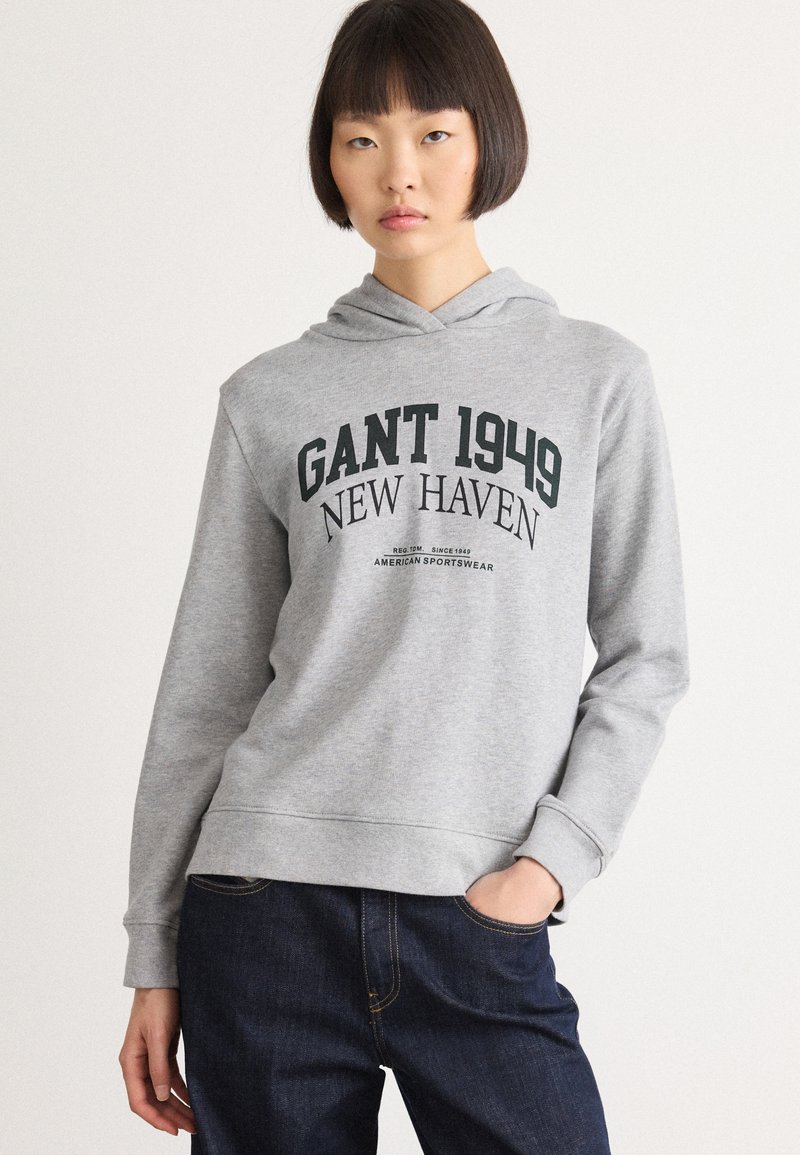 Ung kvinne med bobfrisyre iført en grå hettegenser med teksten "GANT 1949 NEW HAVEN" og mørkeblå jeans, hånd i lommen, nøytral bakgrunn.