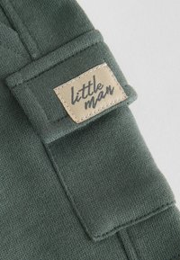 Dunkelgrüner Strickstoff mit einem umgeschlagenen Bund, der ein beiges Etikett mit der Aufschrift "kleiner Mann" in schwarzem Text trägt, mit sichtbaren Fadendetails genäht.