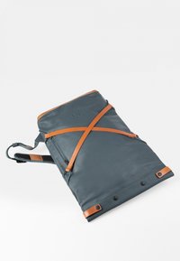 Zaino realizzato in materiale blu scuro con dettagli in pelle marrone. Presenta una tracolla in pelle incrociata e una superficie liscia.