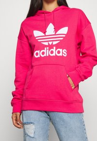 Persona que lleva una sudadera Adidas de color rosa brillante con bolsillo frontal y logo blanco, combinada con unos jeans rotos de color azul claro.