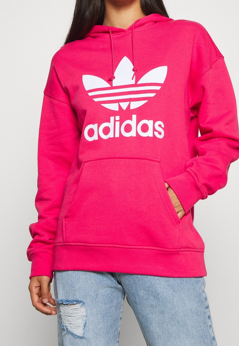 Persona que lleva una sudadera Adidas de color rosa brillante con bolsillo frontal y logo blanco, combinada con unos jeans rotos de color azul claro.