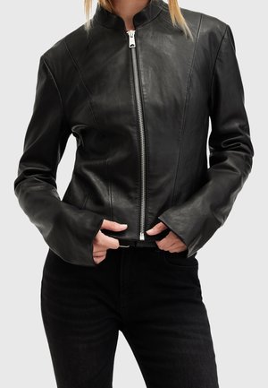 Personne portant une veste en cuir noire ajustée avec une fermeture éclair frontale et des manches longues, portée sur un pantalon noir, sur un fond uni.