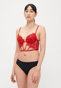 Sutiã vermelho de renda com design estruturado, detalhes florais e painéis transparentes. Apresenta alças ajustáveis e fecho de gancho. Usado com cuecas pretas.