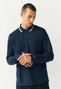 Navyblaues Langarm-Polohemd mit geripptem Kragen und beigen Streifen. Weicher Stoff mit leichter Textur und kleinem gesticktem Logo auf der Brust.