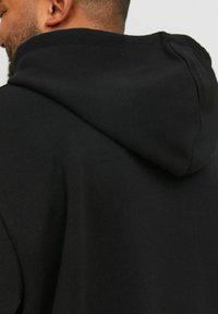 Felpa nera con un tessuto morbido e strutturato. Presenta un grande cappuccio e nessun logo o decorazione visibile. Design semplice e minimalista.