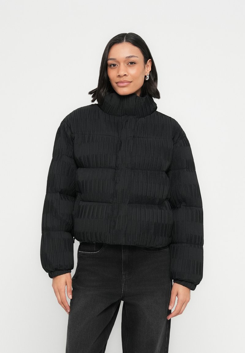 Μαύρο μπουφάν puffer με ψηλό γιακά, ριμπ υφή, κοντό σχέδιο και κούμπωμα με κουμπιά μπροστά. Διαθέτει φαρδιά μανίκια και δεν έχει ορατά λογότυπα.