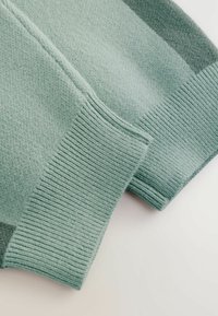 Šviesiai žalias megztuko rankovės galas su įrengta tekstūra, pasižymintis minkšta, megzta medžiaga ir lygia apdaila. Siūlų detalė matoma krašte.