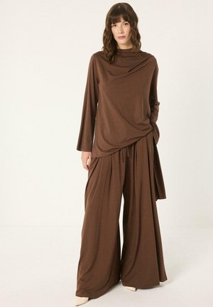 Femme portant un haut marron ample à manches longues et un pantalon à jambes larges, debout sur un fond uni clair, avec une main dans la poche.