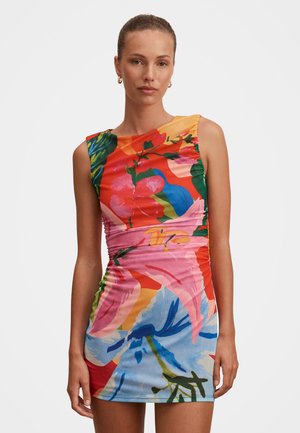 Mujer que lleva un minivestido sin mangas y colorido con estampados florales en verde, rosa, azul, rojo y naranja sobre un fondo liso.