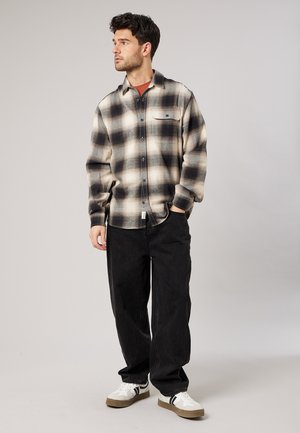 Jonge man staand, draagt een beige en zwarte geruite blouse, zwarte broek, witte sneakers, kijkt naar links tegen een effen grijze achtergrond.