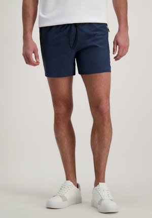 Man draagt marineblauwe shorts met trekkoord, witte sneakers en een wit shirt, staand tegen een effen lichte achtergrond.