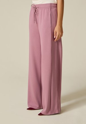 Femme portant un pantalon ample à jambes larges mauve avec taille à cordon, debout devant un fond clair uni, vue de profil de la taille vers le bas.