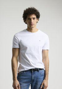 Camiseta blanca de manga corta de algodón, con un escote redondo y un pequeño logo en el lado izquierdo del pecho. Llevada con jeans azules.