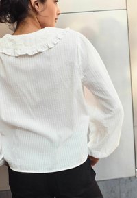 Blouse blanche avec un tissu texturé à motif, dotée d'un col froncé et de manches longues, associée à un pantalon noir.