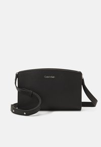Schwarze Leder-Crossbody-Tasche mit strukturierten Oberflächen, Reißverschluss und verstellbarem Tragegurt mit metallischen Akzenten. Markenlogo auf der Vorderseite angezeigt.