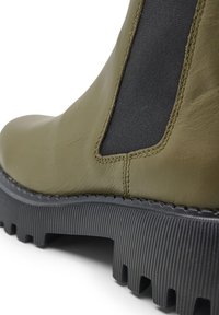 Bottine en cuir vert olive avec des panneaux latéraux élastiques, une semelle en caoutchouc noire texturée et un motif de crampons prononcé.