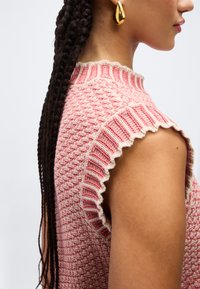 Γυναίκα με μακριά πλεγμένα μαλλιά που φοράει ροζ πλεκτό τοπ χωρίς μανίκια και χρυσό σκουλαρίκι σε σχήμα σταγόνας, εμφανιζόμενη από το πλάι μπροστά σε απλό φόντο.