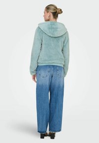 Faux fur hoodie in light blue, featuring a hood and ribbed waistband, paired with loose-fitting blue jeans and heeled shoes.