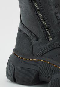 Bottine en cuir noir avec une fermeture éclair sur le côté, une tige texturée et une semelle en caoutchouc épaisse avec des accents de couture jaune.