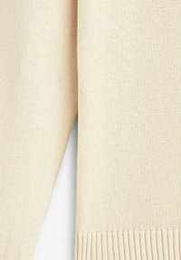 Gros plan sur un tissu tricoté couleur crème avec un ourlet côtelé et un motif de points texturés.