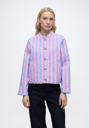 Vrouw draagt een roze en blauw verticaal gestreepte jas met vijf bruine knopen, gecombineerd met donkerblauwe broek, staand tegen een witte achtergrond.