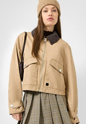 Young woman with long brown hair wearing a beige zip-up jacket, plaid skirt, tan knit hat, and carrying a dark shoulder bag.
