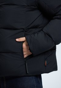 Schwarze Puffjacke mit strukturierter Oberfläche, einer Reißverschlusstasche und elastischen Bündchen, mit einem kleinen Stoffetikett nahe dem Saum.
