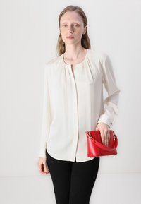 Blusa bianca a maniche lunghe con scollo arricciato e chiusura a bottone, abbinata a una piccola borsa rossa con tracolla corta e dettagli in oro.