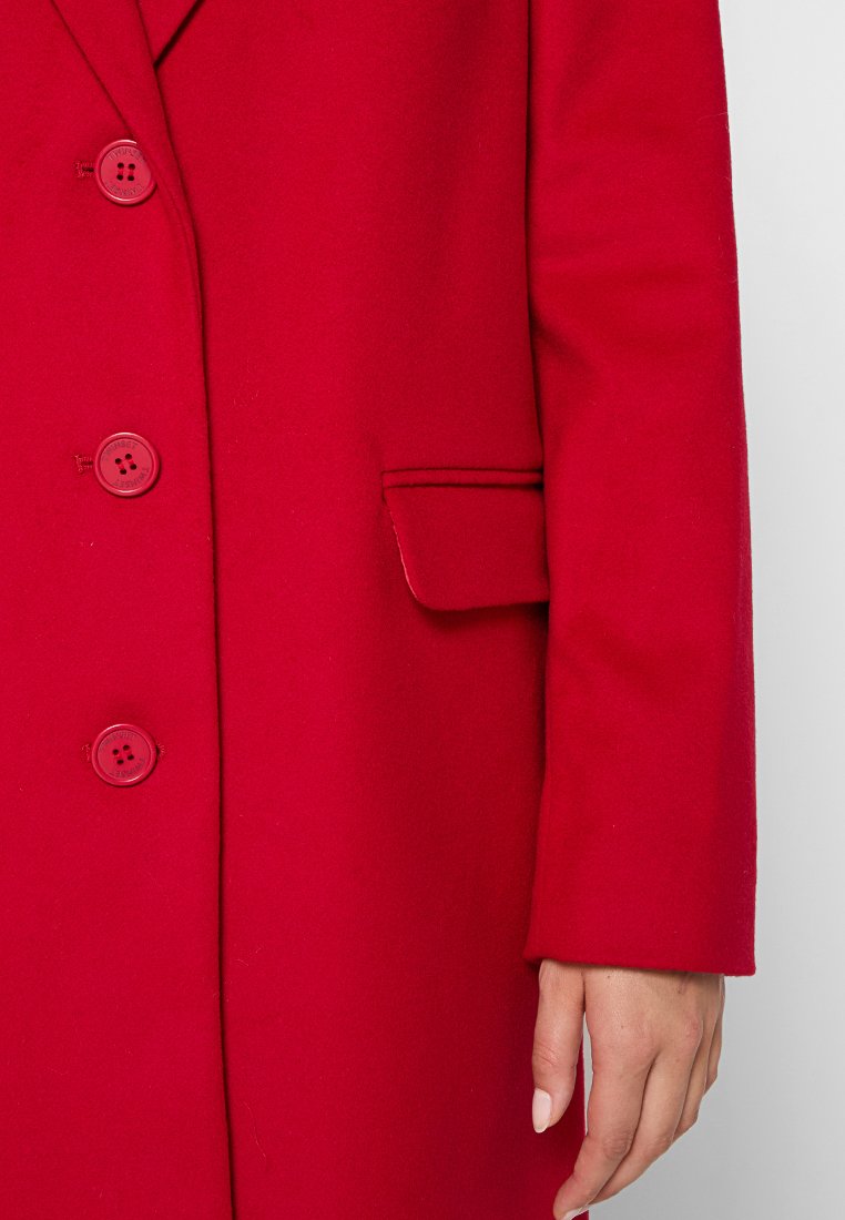Cappotto di lana rosso con tre bottoni rossi, tasca a ribalta e la mano di una persona che riposa accanto alla manica su uno sfondo bianco.