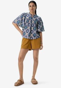 Blouse bleue fluide à motifs avec des manches courtes, associée à un short moutarde et des sandales marron. Présente des imprimés géométriques et une coupe décontractée.